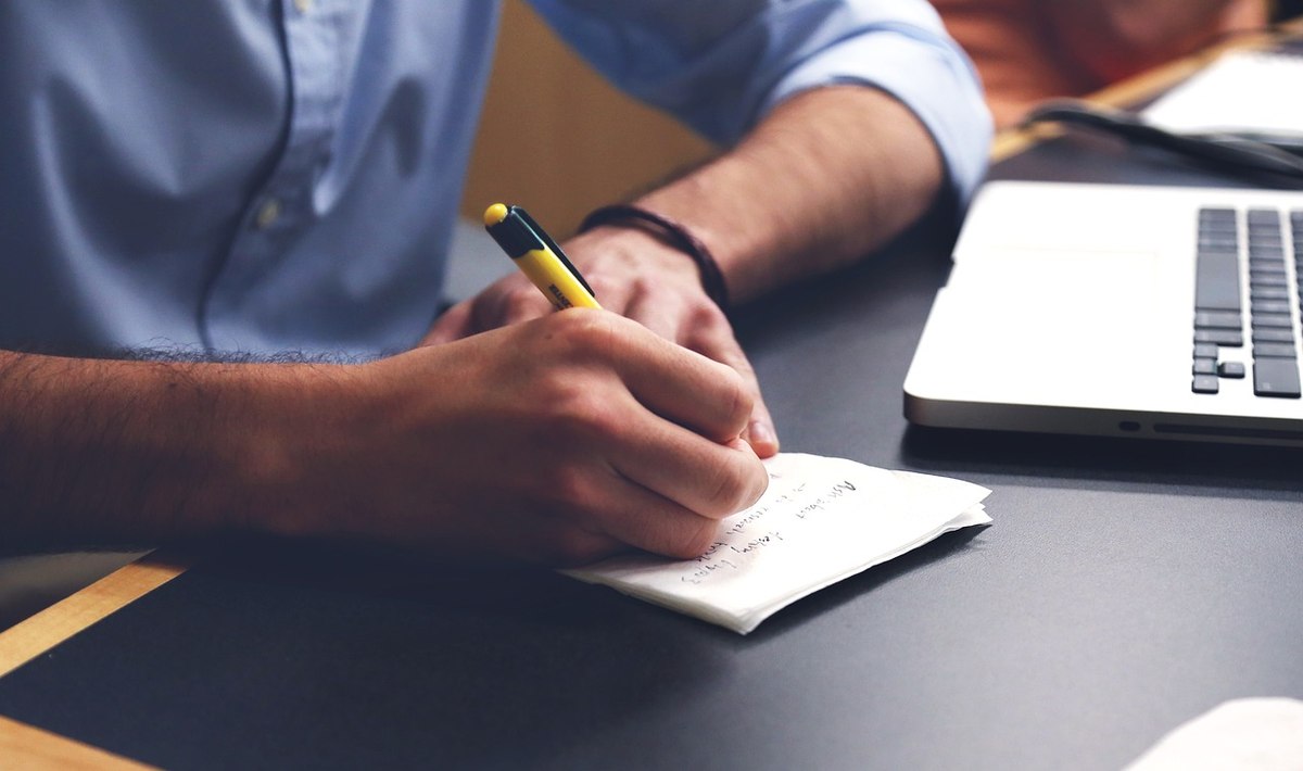 Un homme écrit avec un stylo sur un petit carnet, à côté d'un ordinateur Un homme écrit avec un stylo sur un petit carnet, à côté d'un ordinateur